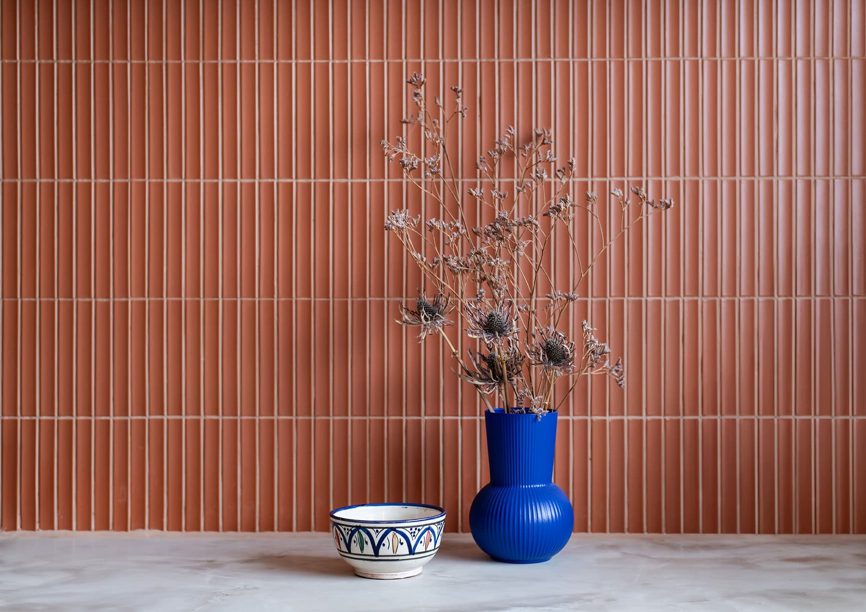 Detaljbilde av flislagt vegg med fingermosaikk i en rusten rødtone. Marmorert benkeplate med en blå vase og dekorert skål